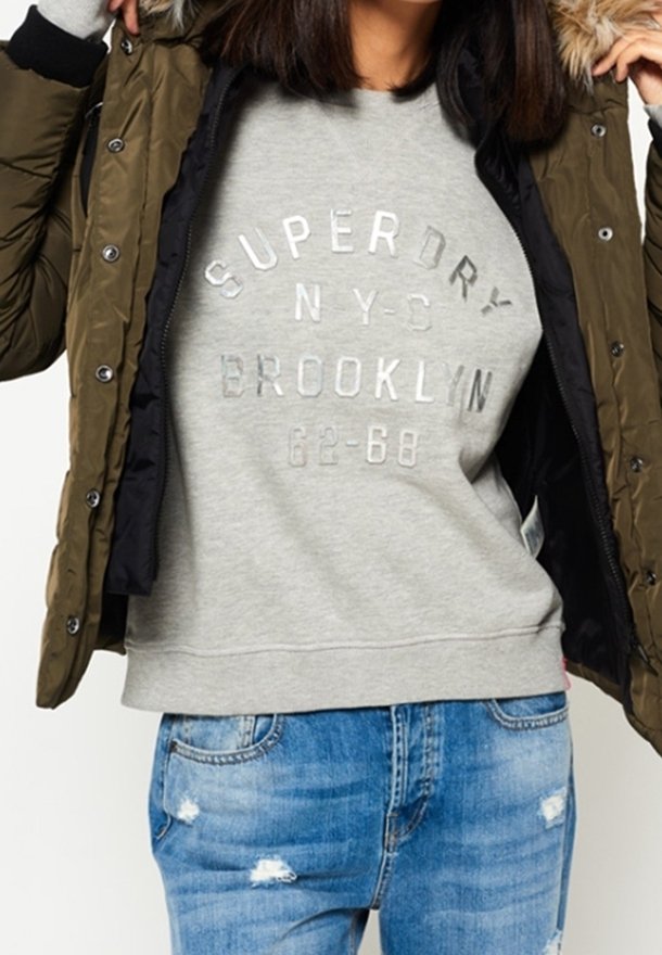 Chaqueta acolchada verde oliva con capucha de piel sintética, combinada con una sudadera gris clara con texto plateado y vaqueros azules desgastados.