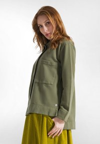 Olijfgroene lange mouwen shirt met een knoopsluiting aan de voorkant en een borstzak. Zachte stof, relaxed fit en zichtbare stikseldetails.