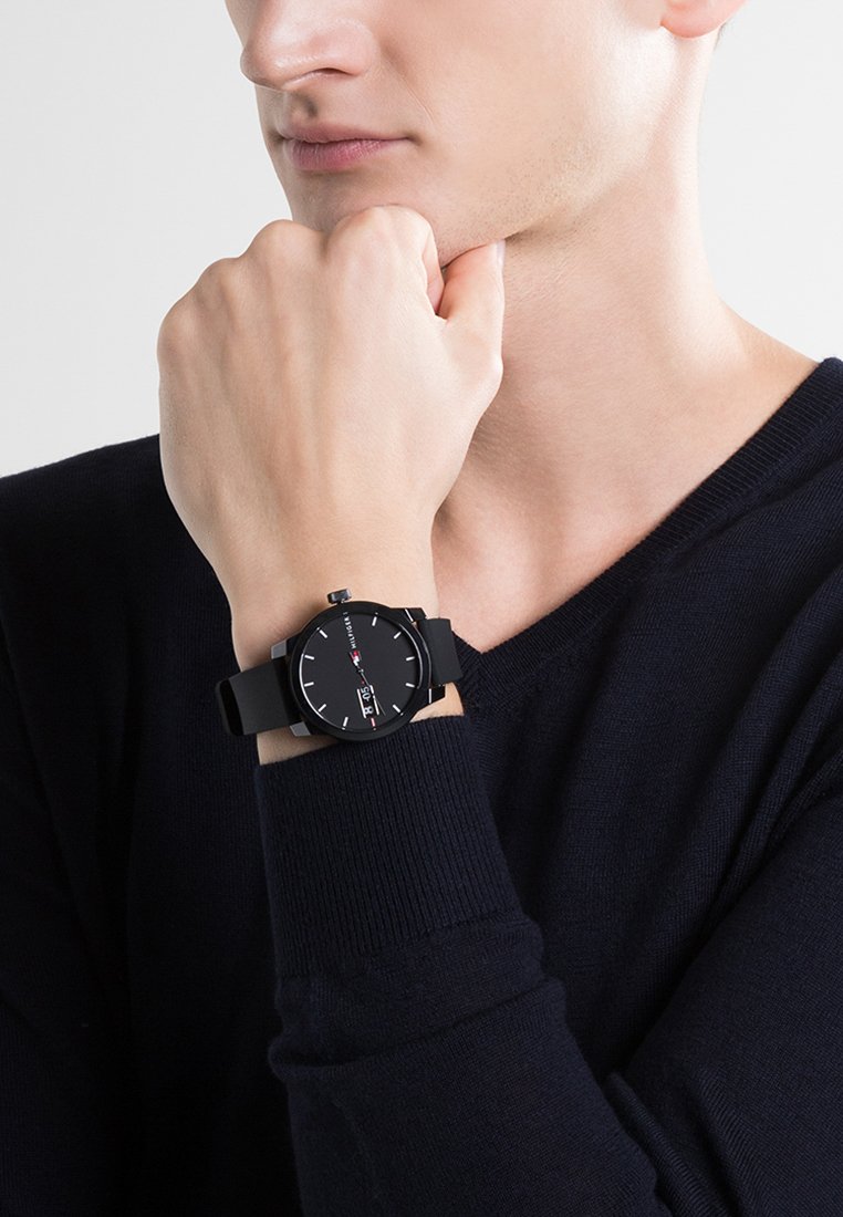 Czarny zegarek na rękę z okrągłą tarczą, minimalistycznym designem, białymi znacznikami godziny i silikonowym paskiem. Noszony na nadgarstku modela.