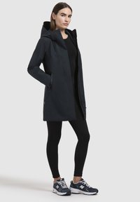 Vattenavvisande marinblå jacka med huva, dragkedjefickor och rak skärning. Bärs över svarta leggings och gråa träningsskor.