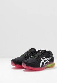 Zapatillas deportivas negras con tejido texturizado, detalles de logo en blanco y una suela translúcida con tonos rosas y verdes.