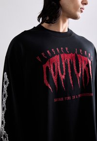 Fekete, hosszú ujjú pólót viselő személy, amelyen piros, studdolt "COUTURE" felirat található, a póló fölött "VERSACE JEANS" szöveggel és lánc grafikákkal az ujjakon.