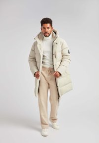 Giacca puffer di un beige chiaro, con cappuccio, design trapuntato e chiusura frontale con zipper. Indossata sopra un dolcevita bianco e pantaloni beige.