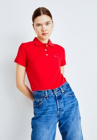 Mujer joven con ojos azules y cabello castaño, vistiendo una camisa polo roja y pantalones vaqueros de tiro alto en un fondo blanco liso.