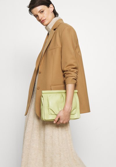 Clutch de couro verde claro com um design franzido e fecho dobrável. O modelo veste um vestido de malha bege e um blazer castanho.