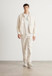 Young man walking indoors wearing cream jacket, white CK logo t-shirt, cream pants, and white sneakers against a plain white wall.