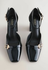 Black patent leather block-heeled shoes with an open back, a square toe, and a decorative gold-tone buckle on the ankle strap.