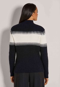 Eine Frau mit langen, dunklen Haaren trägt einen gestreiften dunkelblauen Pullover mit einem weißen horizontalen Streifen und schwarze Hosen, steht vor einem hellgrauen Hintergrund.