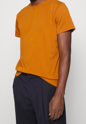 Mand iført en kortærmet orange t-shirt, let stoppet ned i mørk marineblå bukser, står med højre arm afslappet ved siden.