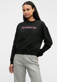 Černá mikina s potiskem "KARL LAGERFELD JEANS" v růžové. Navržena s kulatým výstřihem a žebrovanými manžetami. V kombinaci se šedými kalhotami.