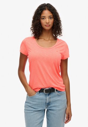 Kobieta o kręconych włosach, ubrana w jasnoróżowy krótki rękaw, włożona w jasnoniebieskie dżinsy z czarnym paskiem, stoi z jedną ręką w kieszeni.