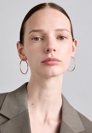 Guldtonede hoops øreringe med en tekstureret overflade, båret af en model iført en afdæmpet brun blazer med en lige halsudskæring og naturlig makeup.