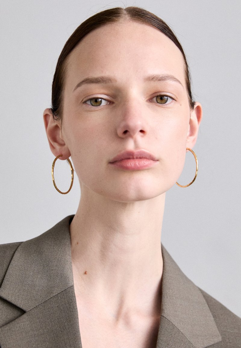 Cercei cerc cu ton de aur, cu o suprafață texturată, purtați de un model într-un sacou maro mat, având un decolteu drept și un machiaj natural.