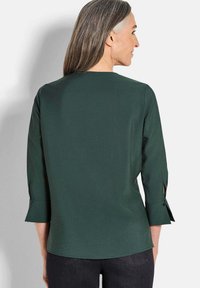 Dunkelgrünes, leichtes Blusenshirt mit Drei-Viertel-Ärmeln, versehen mit Knopfdetails an den Bündchen, glatter Textur und einem lockeren Schnitt.