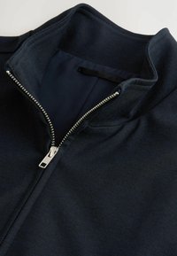 Dunkelblaue Zip-Up-Jacke mit hohem Kragen, glatter Gewebestruktur und einem metallischen Reißverschluss mit stylischem Ziehband.