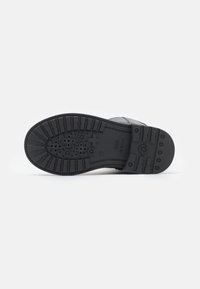 Semelle de chaussure en caoutchouc noir avec un motif de semelle texturé et des trous de drainage, présentant un logo en relief et des caractéristiques de traction le long des bords.