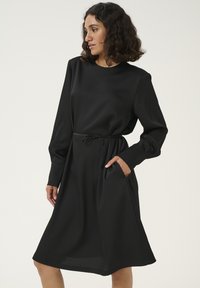 Robe noire à mi-longueur avec manches longues, encolure bateau, taille avec cordon de serrage et poches latérales. Fabriquée en tissu lisse avec une texture subtile.