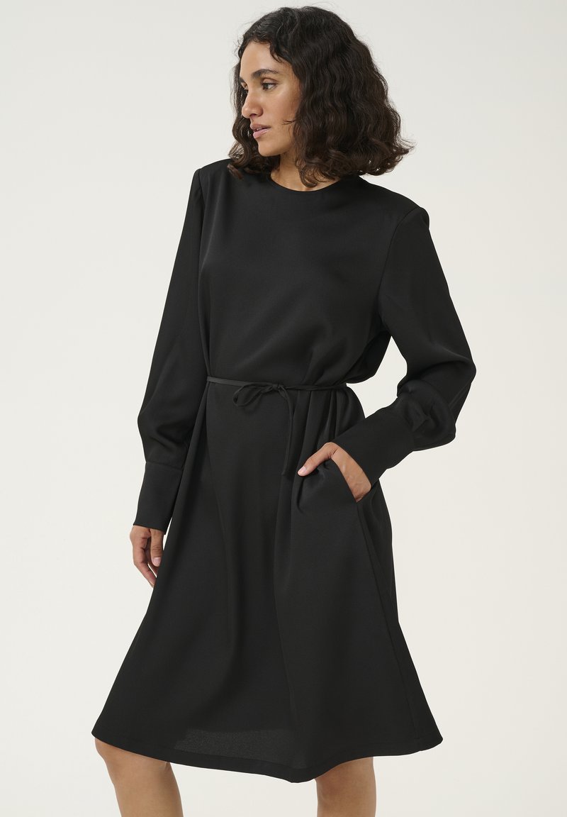Robe noire à mi-longueur avec manches longues, encolure bateau, taille avec cordon de serrage et poches latérales. Fabriquée en tissu lisse avec une texture subtile.