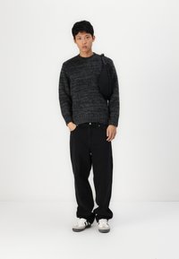 Pull en tricot gris foncé avec un motif texturé, pantalon noir ample, et baskets blanches avec des accents gris et noirs, plus un petit sac noir.
