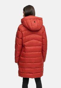 Kvinna iklädd en knälång, röd vadderad jacka med huva, stående vänd bort från kameran mot en enkel ljus bakgrund.