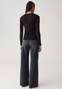 Femme aux longs cheveux bruns, portant un haut noir à manches longues côtelé et un pantalon large en jean noir délavé, de dos contre un fond uni.