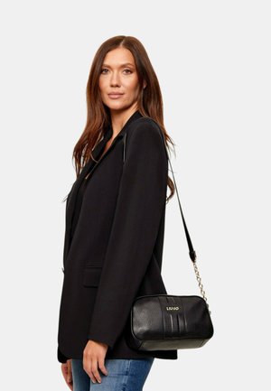 Donna con lunghi capelli castani che indossa un blazer nero e jeans blu, con una borsa a tracolla nera Liu Jo sulla spalla.