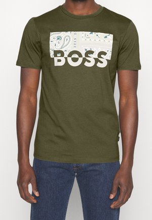 Mand iført olivengrøn kortærmet t-shirt med blokmønstret design og store hvide bogstaver, der stavner "BOSS" på forsiden, kombineret med blå jeans.