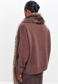 Brązowy sweter dzianinowy z kołnierzem z sztucznego futra, o luźnym kroju, ze ściągaczem u dołu i miękkiej teksturze, noszony na brązowej spódnicy.