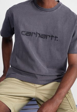 Mand iført en falmet mørkegrå Carhartt T-shirt og beige shorts, siddende med hænderne hvilende på lårene.