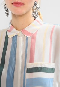 Blouse rayée multicolore avec un col boutonné, une poche avant avec une rayure horizontale verte et bleue, et un tissu léger.