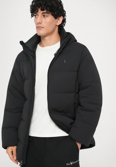 Mladenič oblečen v črno puhovko preko bele srajce in črne trenirke Polo Ralph Lauren, ki gleda levo proti enotnemu ozadju.