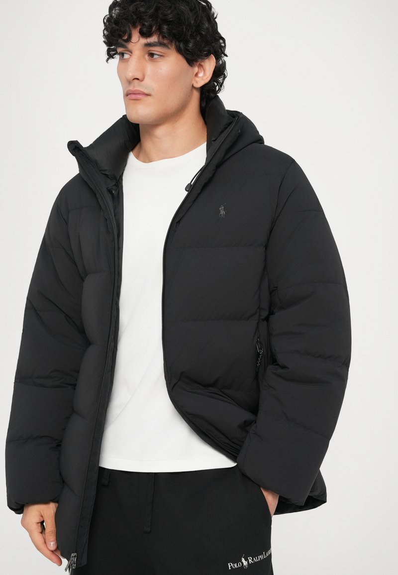 Joven vestido con una chaqueta de plumas negra sobre una camiseta blanca y pantalones de chándal negros de Polo Ralph Lauren, mirando a la izquierda contra un fondo simple.