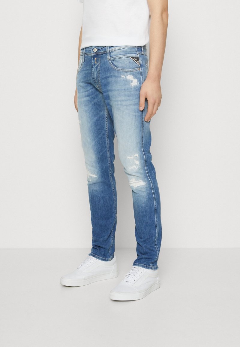 Personne portant un jean slim bleu clair délavé avec des zones usées, des baskets blanches et un t-shirt blanc uni à manches courtes.