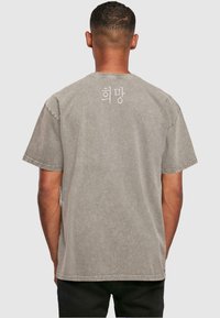Camiseta gris de manga corta hecha de algodón con un aspecto desgastado. Presenta un diseño de texto en coreano en la parte superior de la espalda y detalles cosidos.