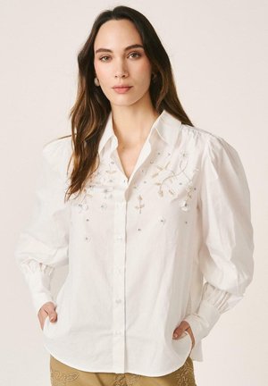 Mujer con cabello largo y castaño, vistiendo una camisa blanca abotonada con mangas abullonadas y delicados bordados florales, con las manos en los bolsillos, fondo neutro.