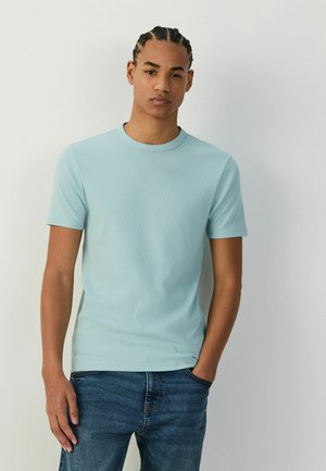 Young man with braided hair wearing a light blue short-sleeve shirt and blue jeans, standing against a plain light background.