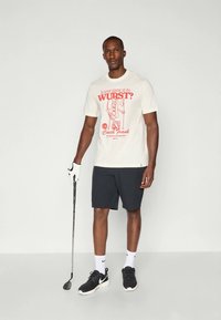Férfi fehér grafikás pólóban, fekete rövidnadrágban és sportcipőben, aki golfütőt tart a kezében, egy sima világos háttér előtt áll.