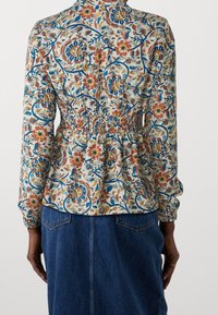 Blusa a maniche lunghe con motivo floreale, vita arricciata, sfondo crema chiaro e accenti vivaci di blu, rosso e giallo.