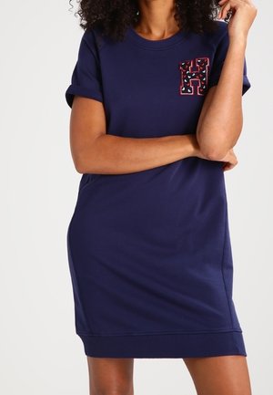 Femme portant une robe bleu marine à manches courtes avec un patch "H" à motifs sur la poitrine, debout, les bras croisés.