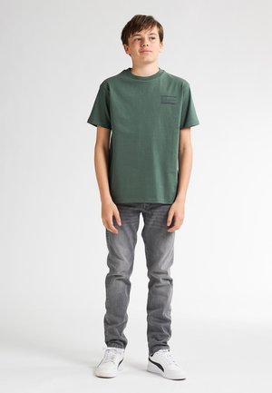 T-shirt verde in cotone con maniche corte, collezione a girocollo e dettaglio stampato sul petto, abbinata a jeans grigi e sneaker bianche.
