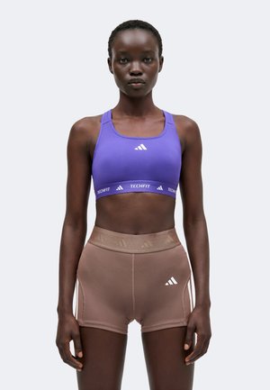 Žena s fialovou športovou podprsenkou Adidas a hnedými vysokými šortkami Adidas stojí na jednofarebnom svetlom pozadí.