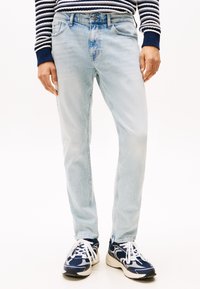 Jeans din denim albastru deschis cu tăietură dreaptă, design cu cinci buzunare și ușoară decolorare, asociate cu adidași atletici negri și albi.