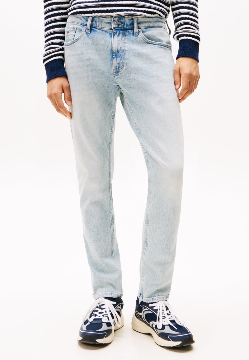 Jeans din denim albastru deschis cu tăietură dreaptă, design cu cinci buzunare și ușoară decolorare, asociate cu adidași atletici negri și albi.