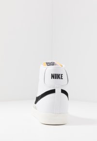 Weiße High-Top-Sneaker mit schwarzen Akzenten, Obermaterial aus Leder, gepolsterter Kragen und strukturierten Außensohle. Merkmale des markanten "NIKE"-Logos an der Ferse.