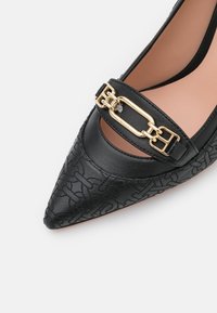 Chaussure plate noire pointue avec motif embossé et détail de boucle chaine dorée sur la partie supérieure.