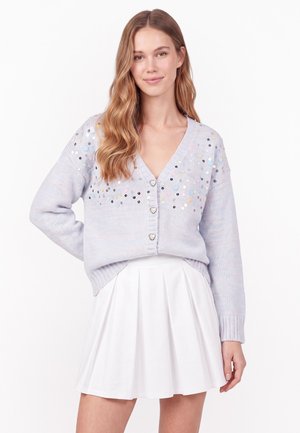 Jeune femme aux longs cheveux ondulés portant un cardigan violet clair avec des sequins et des boutons en forme de cœur et une jupe plissée blanche.