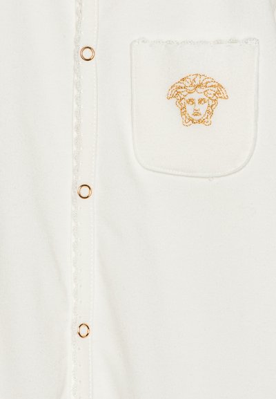 Camisa blanca con bordado dorado de una cara en el bolsillo, con acentos de botones dorados y un detalle de tejido texturizado a lo largo de la tapeta.