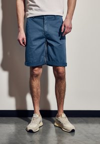 Modré bavlněné šortky s ohrnutými lemy, mají rovný střih a přední kapsy, doplněné o světlé šedé tenisky na betonové podlaze.