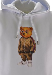 Felpa bianca con una stampa di un orso con occhiali, tuta marrone con motivo e strisce rosse e blu, un orologio da polso e sneakers bianche.