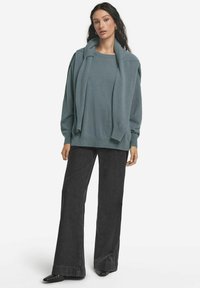 Överdimensionerad tealtröja i lager över en matchande topp, kombinerad med svarta vida jeans och spetsiga svarta skor. Slät tyg med ribbade detaljer.